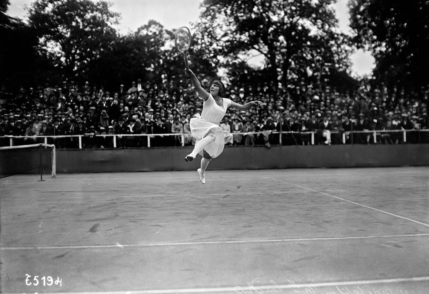 Suzanne Lenglen saltando para uma bola, com as arquibancadas lotadas ao fundo