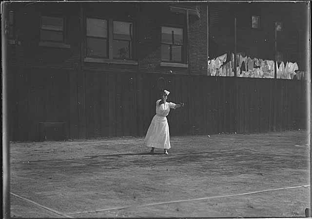 Mulher jogando tennis por volta de 1910, de vestido longo branco até o tornozelo, colarinho alto e chapéu